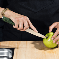 A person cutting a green apple with a knife on a wooden cutting board.